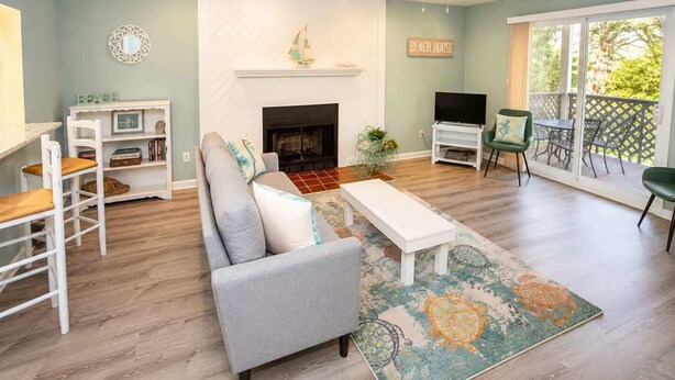 A living room with a fireplace, a TV, a sofa, and a balcony with outdoor furniture. - 2BR Beach Bungalow | Walk to Beach | Sleeps 5 (Norfolk)