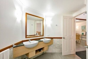 Un baño con dos lavamanos, un espejo y una puerta que da a otra habitación.