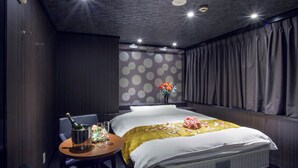 Quarto de hotel com uma cama, uma mesinha redonda com uma garrafa e copos, um vaso com flores e uma parede com bolinhas.