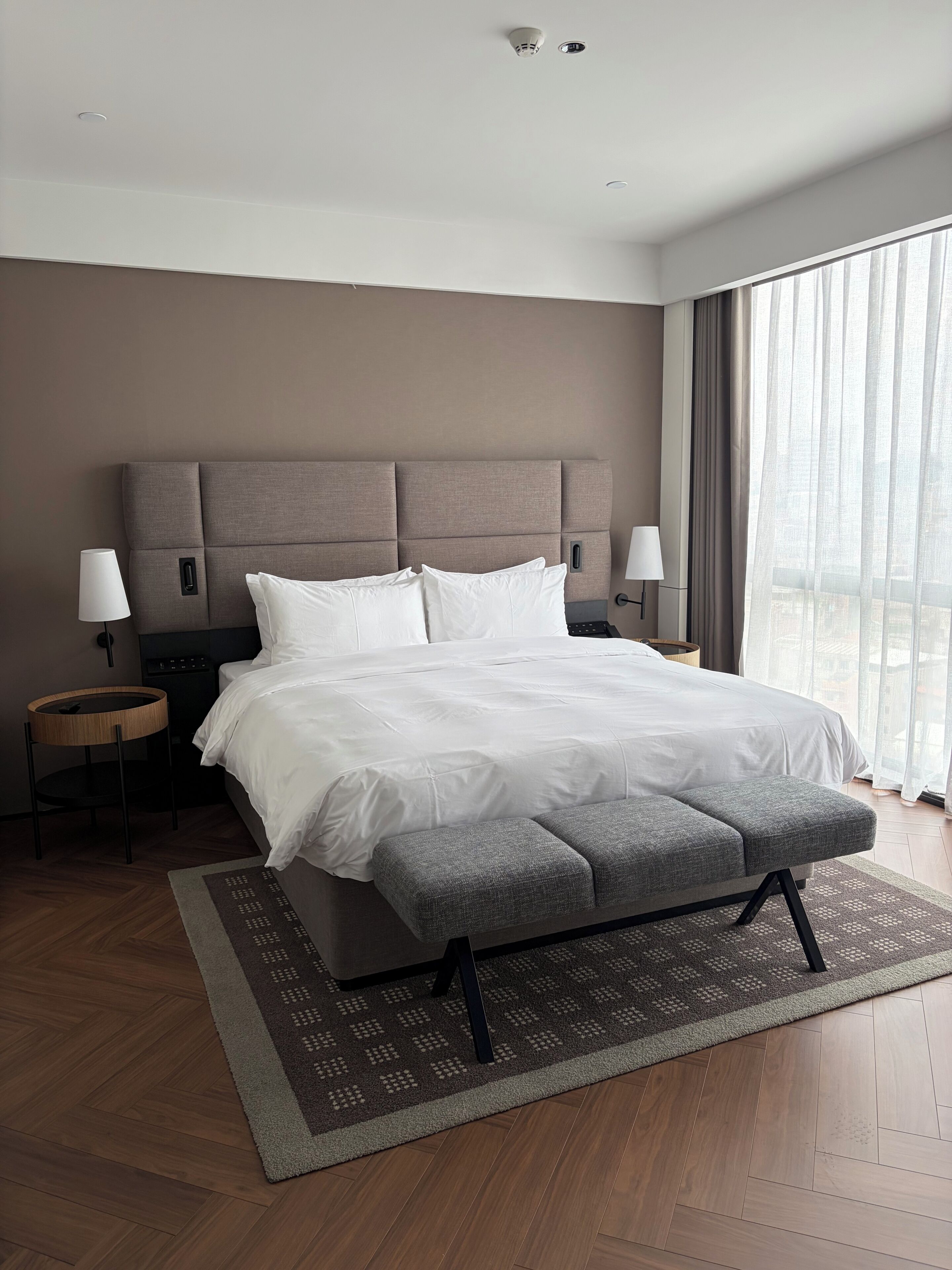 Une chambre d’hôtel moderne, dotée d’un grand lit, de deux tables de chevet, d’un banc et offrant une vue sur la ville grâce à de grandes fenêtres.