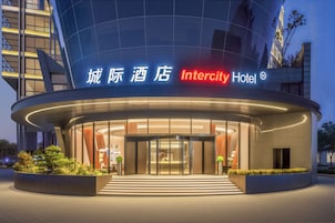 A modern hotel entrance with a glass facade, signage in multiple languages, and a landscaped area with plants.