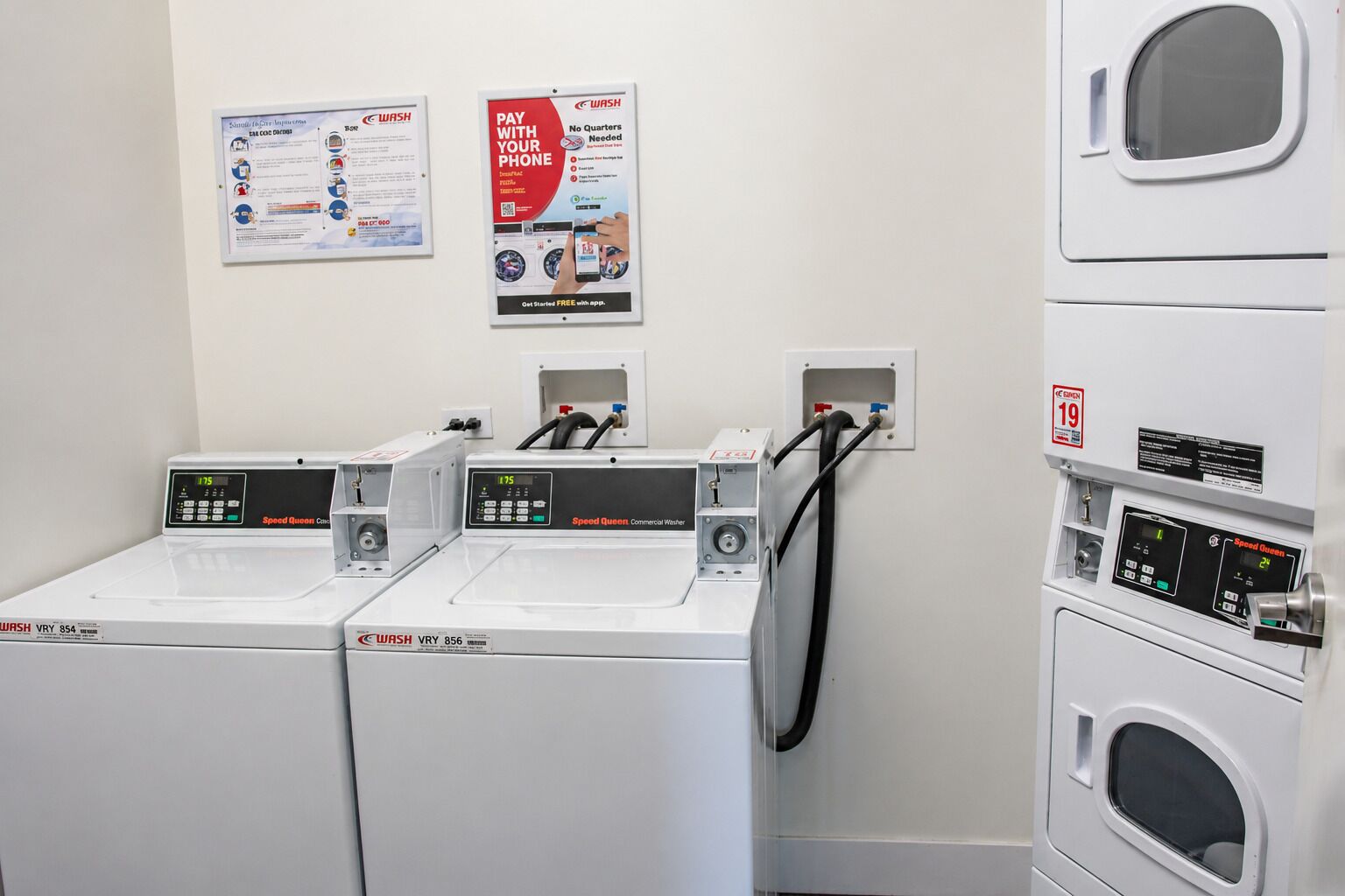 A laundry facility with three WASH brand machines, a poster advertising a mobile payment service, and a safety sign.
