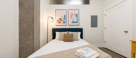 A bedroom with a bed, a headboard, a wall-mounted light, two framed artworks, a door, and a small wooden table.