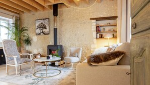 A cozy living room with a stone wall, a fireplace, a sofa with a fur throw, a patterned armchair, and a small table with a lamp.