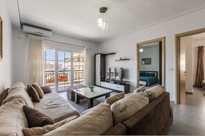 A modern living room with a sofa, coffee table, TV, and a balcony with a view. - Flower Balcony Apartment 2BR Beachside Escape (Sarandë)