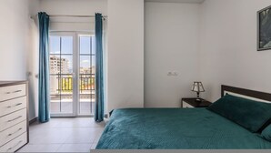 Een slaapkamer met een bed, een nachtkastje, een kapstok, een raam met gordijnen en uitzicht op gebouwen.