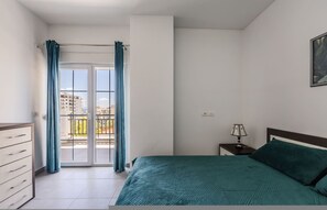 A bedroom with a bed, a nightstand, a dresser, a window with curtains, and a view of buildings outside. - Flower Balcony Apartment 2BR Beachside Escape (Sarandë)
