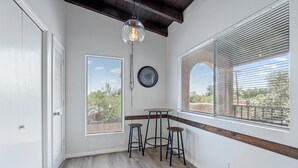 Una habitación con techo de madera, una mesa redonda con taburetes, un reloj de pared y ventanas grandes con persianas.