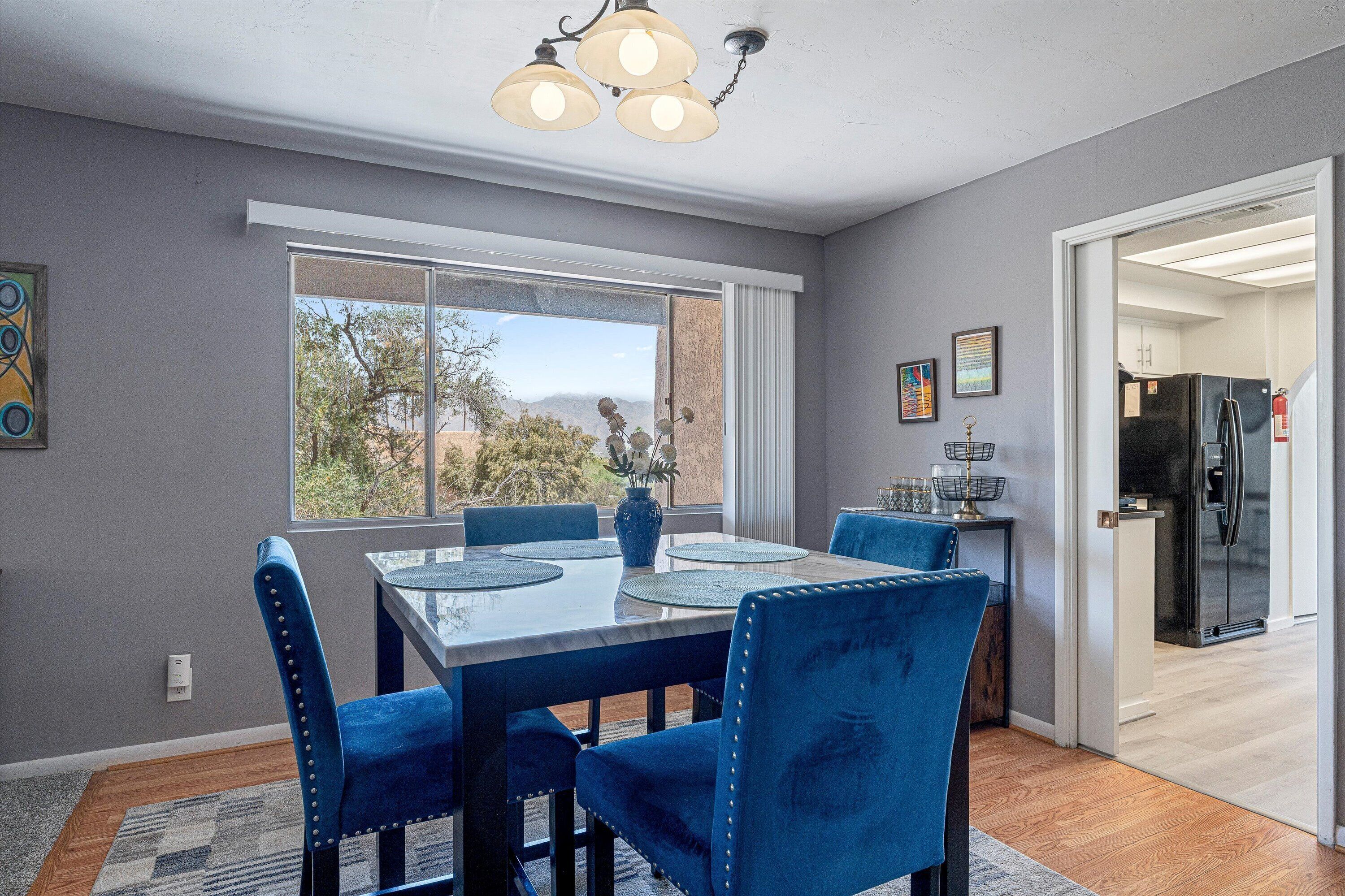 A dining area with a table and chairs, a window with a view, and a kitchen in the background.