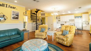 A living room with a sofa, two armchairs, a coffee table, a patterned ottoman, a staircase, and a kitchen area. - Ocean View l Big 5BR Home l Easy Beach Access (Surfside Beach)