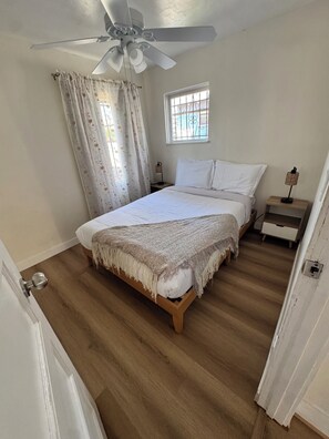 Ein Schlafzimmer mit Bett, Deckenventilator, Fenster mit Gitterstäben, Nachttisch und Beistelltisch.