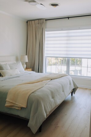 Un dormitorio con cama, mesita de noche, lámpara, ventana con cortinas y vistas al exterior.