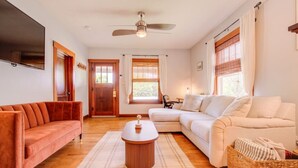 A living room with a sofa, a coffee table, a TV mounted on the wall, and a ceiling fan. - Charming Bungalow 4bd/2ba, Huge Deck and Sunroom (Manhattan)