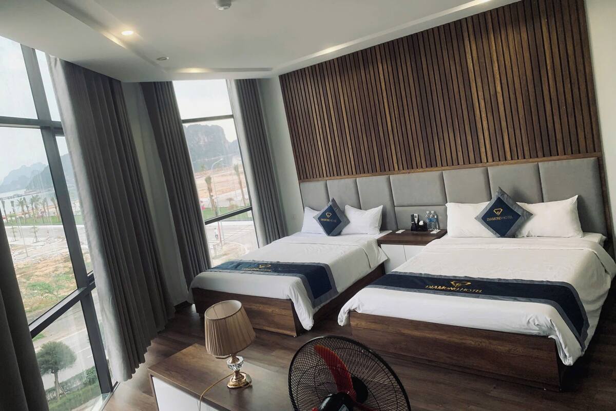 A hotel room with two beds, a desk, a lamp, a fan, and a view of the outdoors.