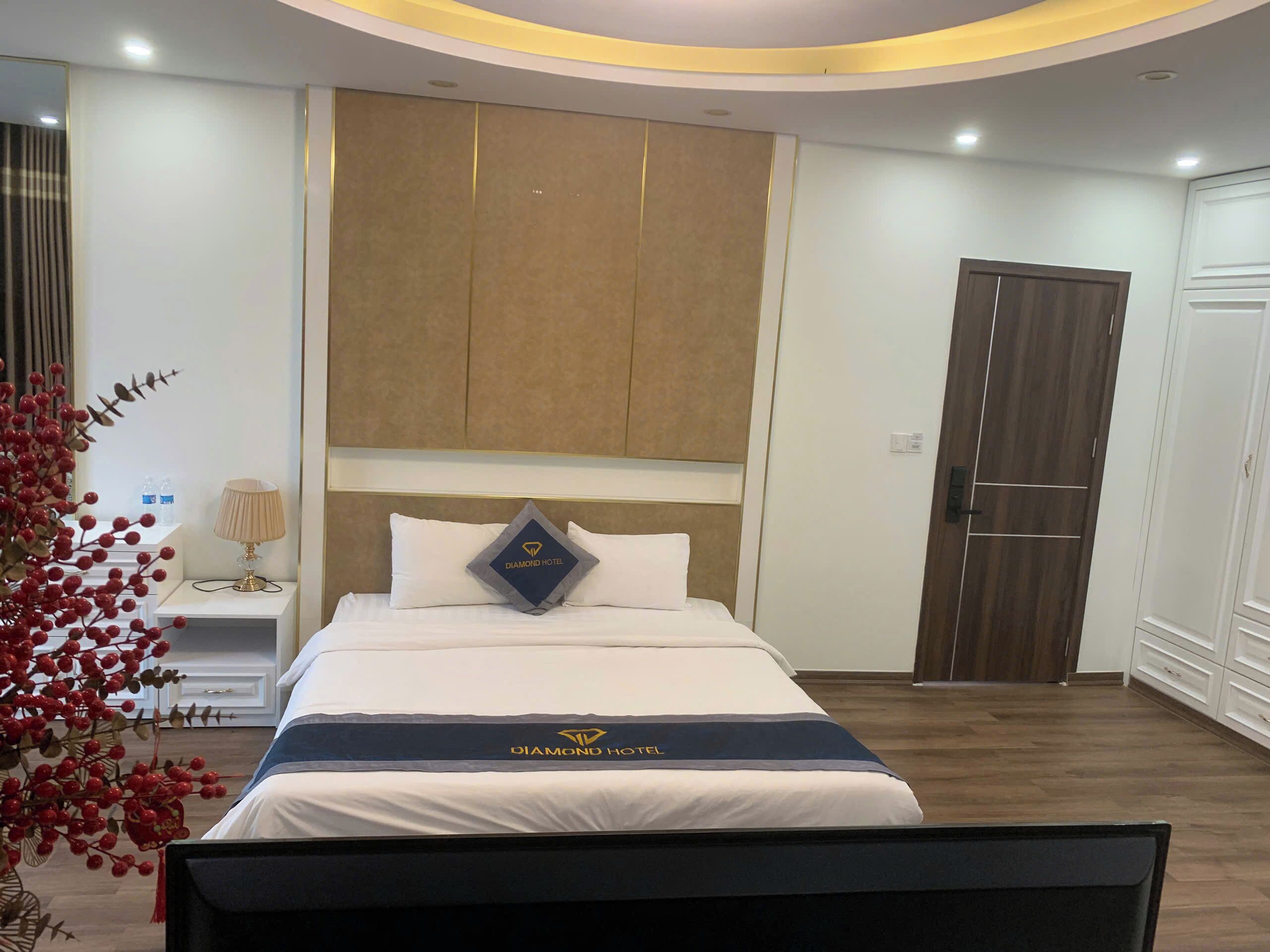 Habitación de hotel con cama, mesita de noche, lámpara, planta decorativa y armario.