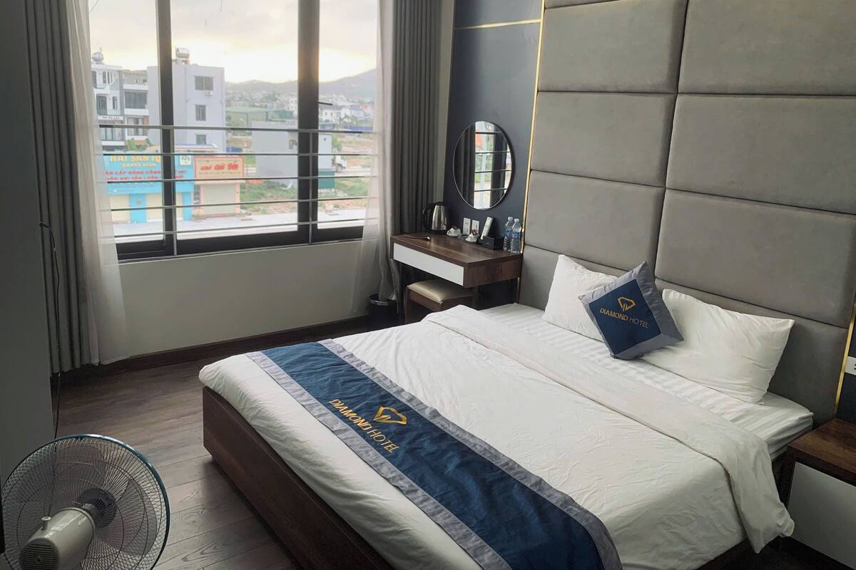 A hotel room with a bed, a bedside table, a mirror, a window with a view of buildings, and a fan.