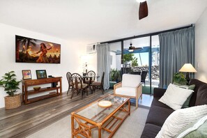 A living room with a sofa, a glass coffee table, a dining area with chairs, a wooden console table, and a painting on the wall. - 🏖️Kona Condo with AC | Walk to Honl's Beach + Partial Ocean View (Kailua Kona)
