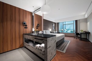 A modern hotel room with a wooden wall, a large bed, a built-in vanity, and a large window offering a city view.