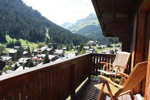 Ein Holzbalkon mit zwei Holzstühlen, mit Blick auf ein dörfliches Bergpanorama mit Häusern und blauem Himmel.