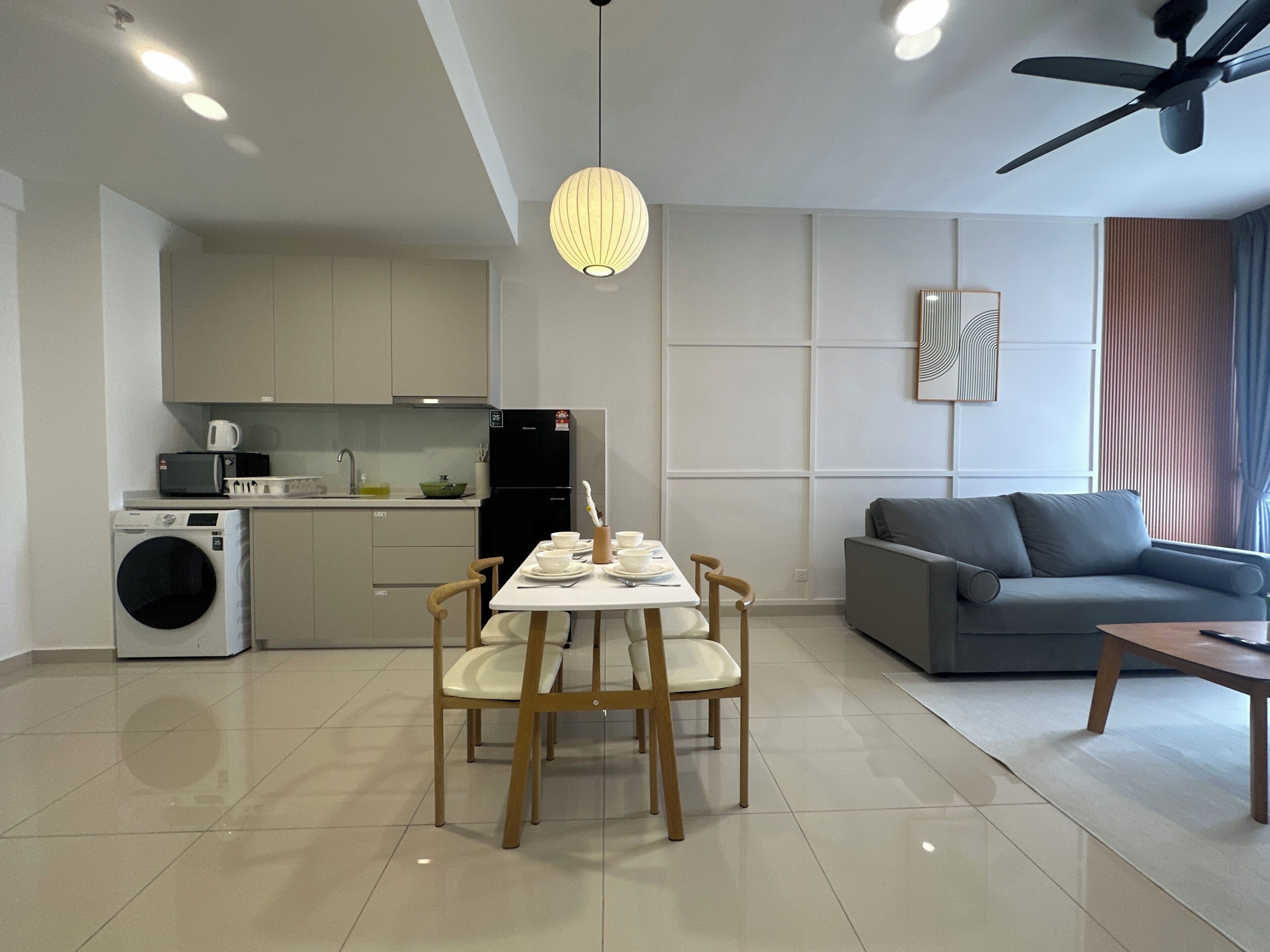 A modern kitchen with a microwave, refrigerator, and washing machine. There is a dining area with a table and chairs, and a gray sofa in the living room.