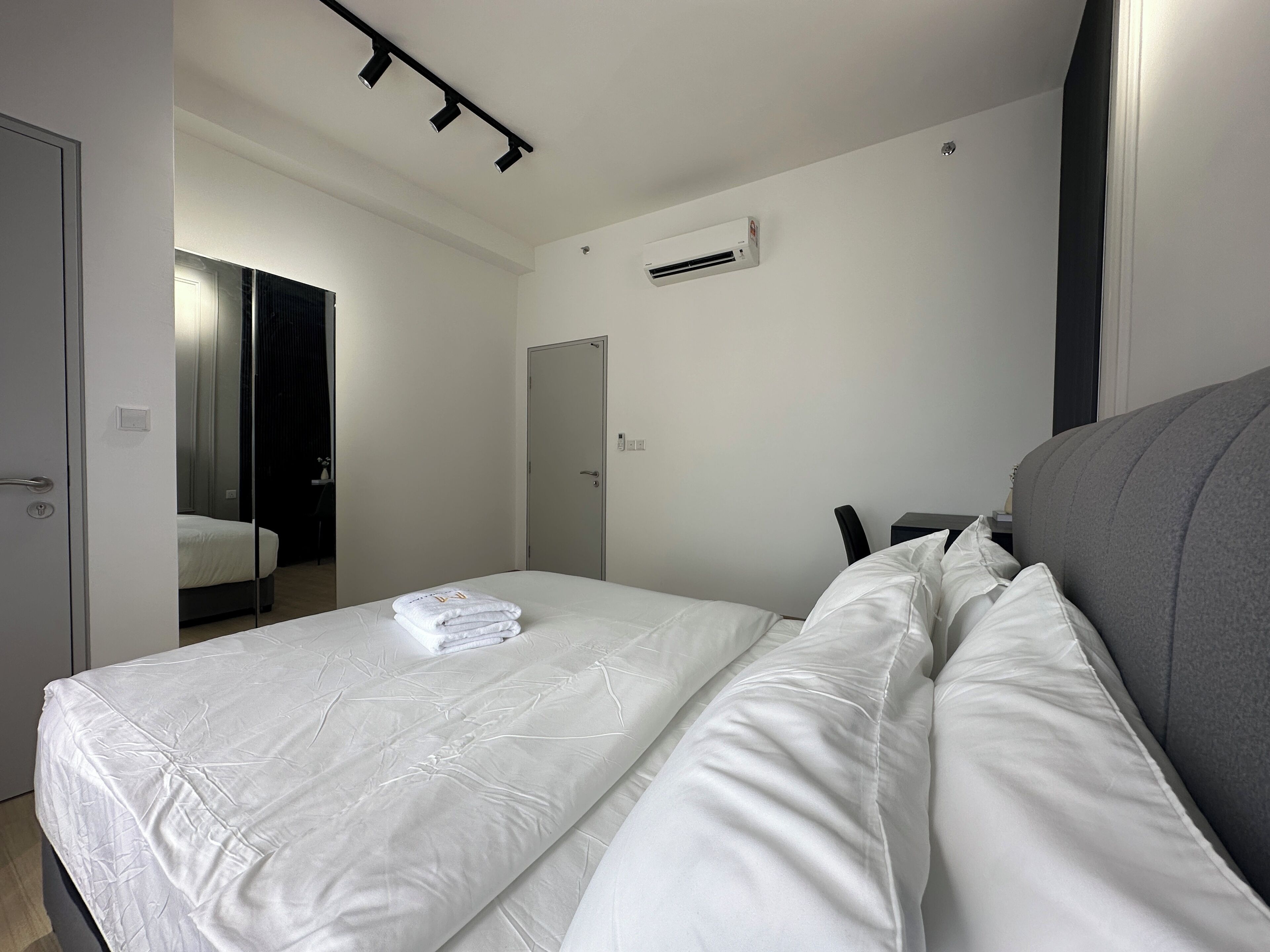 A hotel room with a bed, a desk, a chair, a mirror, and an air conditioning unit.