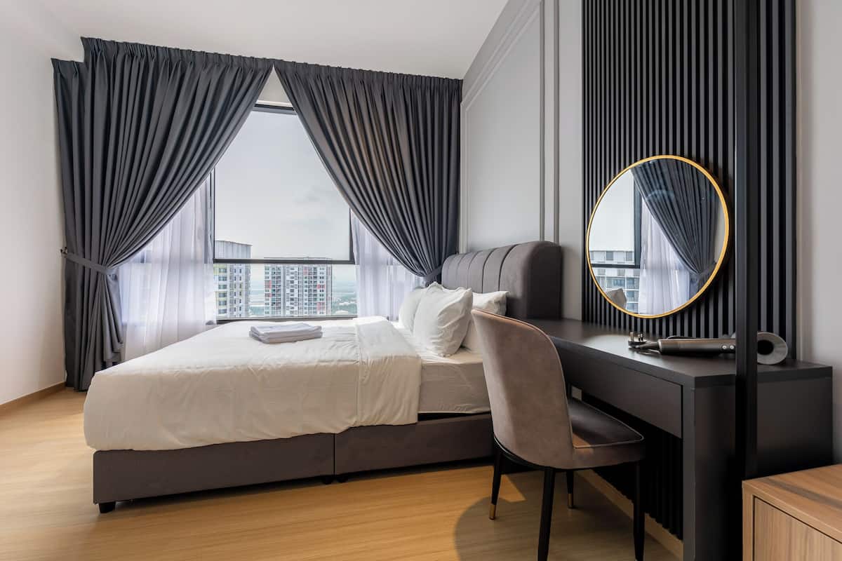 Un dormitorio moderno con cama, escritorio, silla, espejo redondo y vistas de la ciudad a través de amplias ventanas.