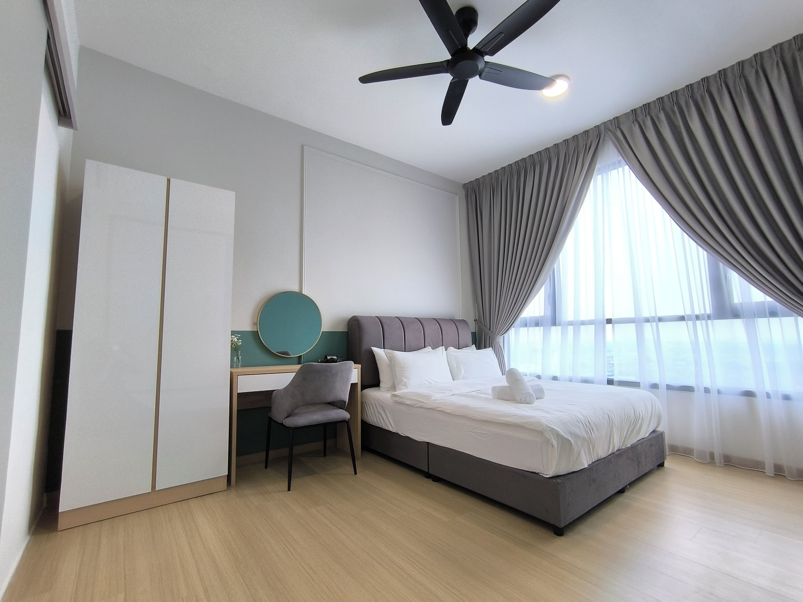 A hotel room with a bed, a desk, a chair, a ceiling fan, and a large window with curtains.