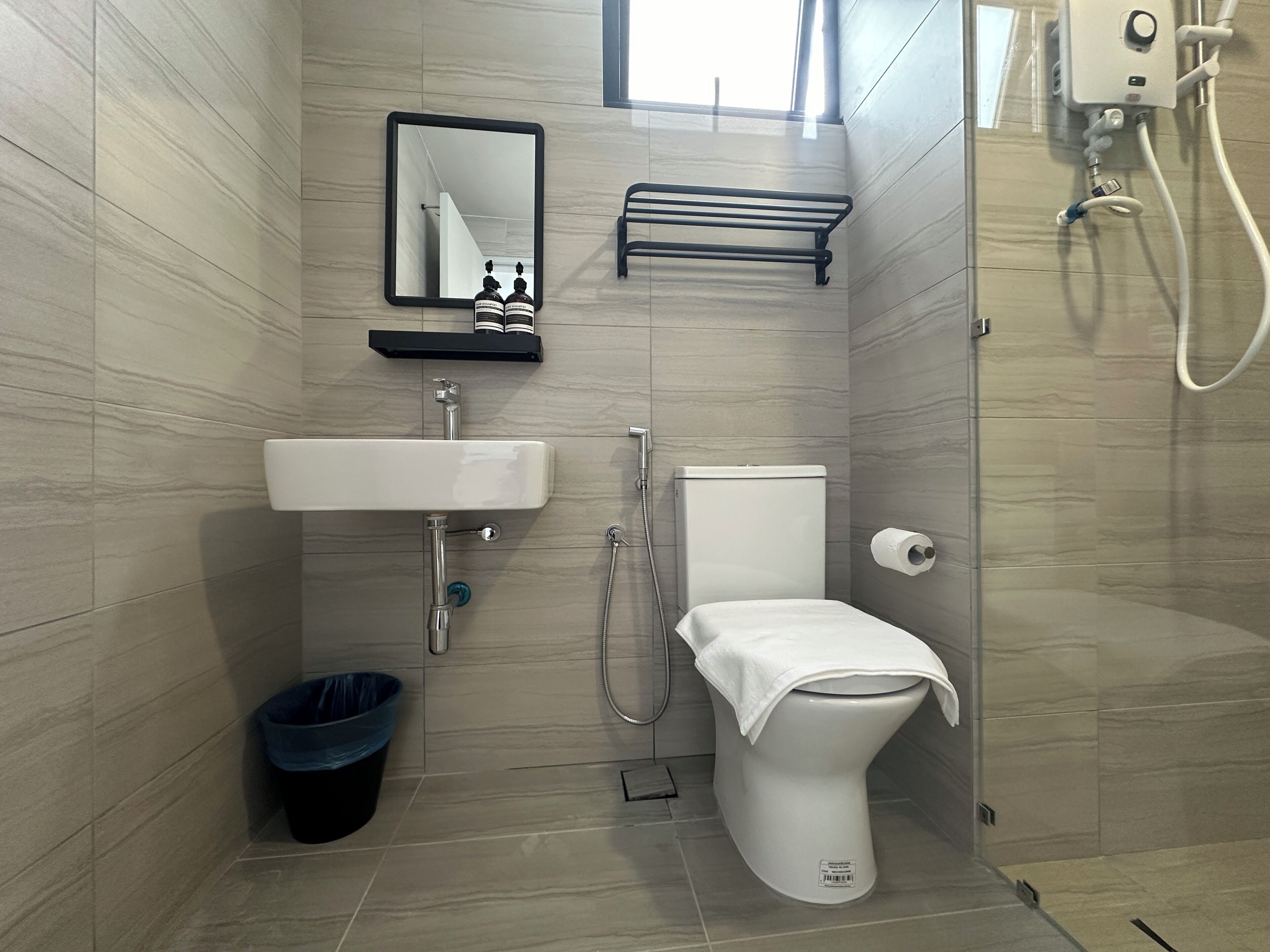 Een badkamer met een wit wastafel, toilet en douche. Er staat een zwarte prullenbak en er is een handdoekhouder.