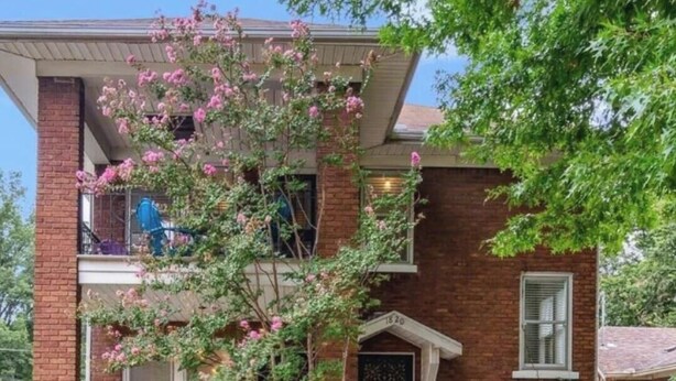 Una casa de ladrillo de dos pisos con un porche delantero, un jardín pequeño y un árbol con flores rosadas.