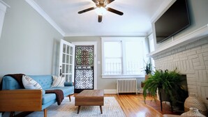 Una sala de estar con un sofá azul, una mesa de centro de madera, un ventilador de techo, una chimenea y un televisor montado en la pared.