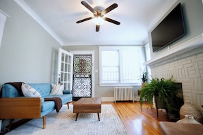 A living room with a blue sofa, a wooden coffee table, a ceiling fan, a fireplace, and a TV mounted on the wall. - Charming Midtown home in the heart of Memphis (Memphis)