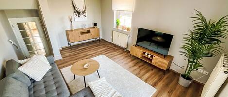 Una sala de estar con un sofá gris, una mesa de centro, un televisor sobre un mueble de madera, un radiador y una planta en maceta.