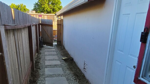 A narrow pathway with stepping stones leading to a white door, flanked by a wooden fence and a building with a red trim.