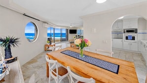 Una cucina moderna con mobili bianchi, una zona pranzo con tavolo e sedie in legno, e vista sull'oceano attraverso un'ampia finestra.