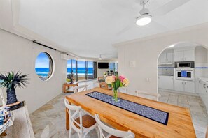 Eine moderne Küche mit weißen Schränken, ein Essbereich mit einem Holztisch und Stühlen sowie Blick auf den Ozean durch ein großes Fenster.