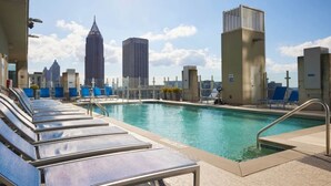 Ein Pool auf dem Dach mit Liegestühlen, im Hintergrund eine Stadtansicht und ein klarer Himmel.