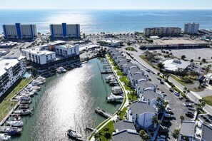An aerial view of a marina with boats docked, a road running parallel to the water, and buildings with balconies overlooking the marina. - Beautifully Updated Madeira Beach Yacht Club Condo | Heated Pool | King Bed | Walk to the Beach (Madeira Beach)