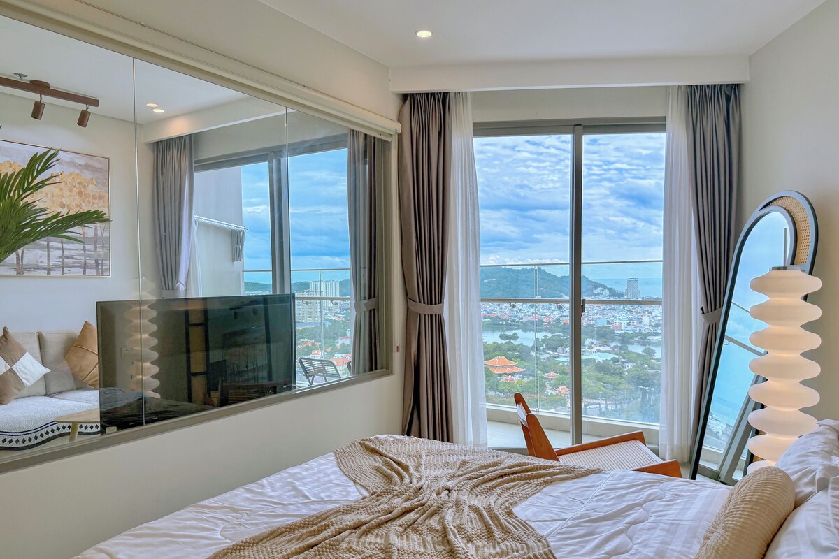 A hotel room with a bed, a TV, a large window with curtains, and a view of a cityscape.