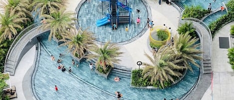 Una gran piscina curvada con múltiples carriles y un tobogán en el centro, rodeada de palmeras y vegetación.