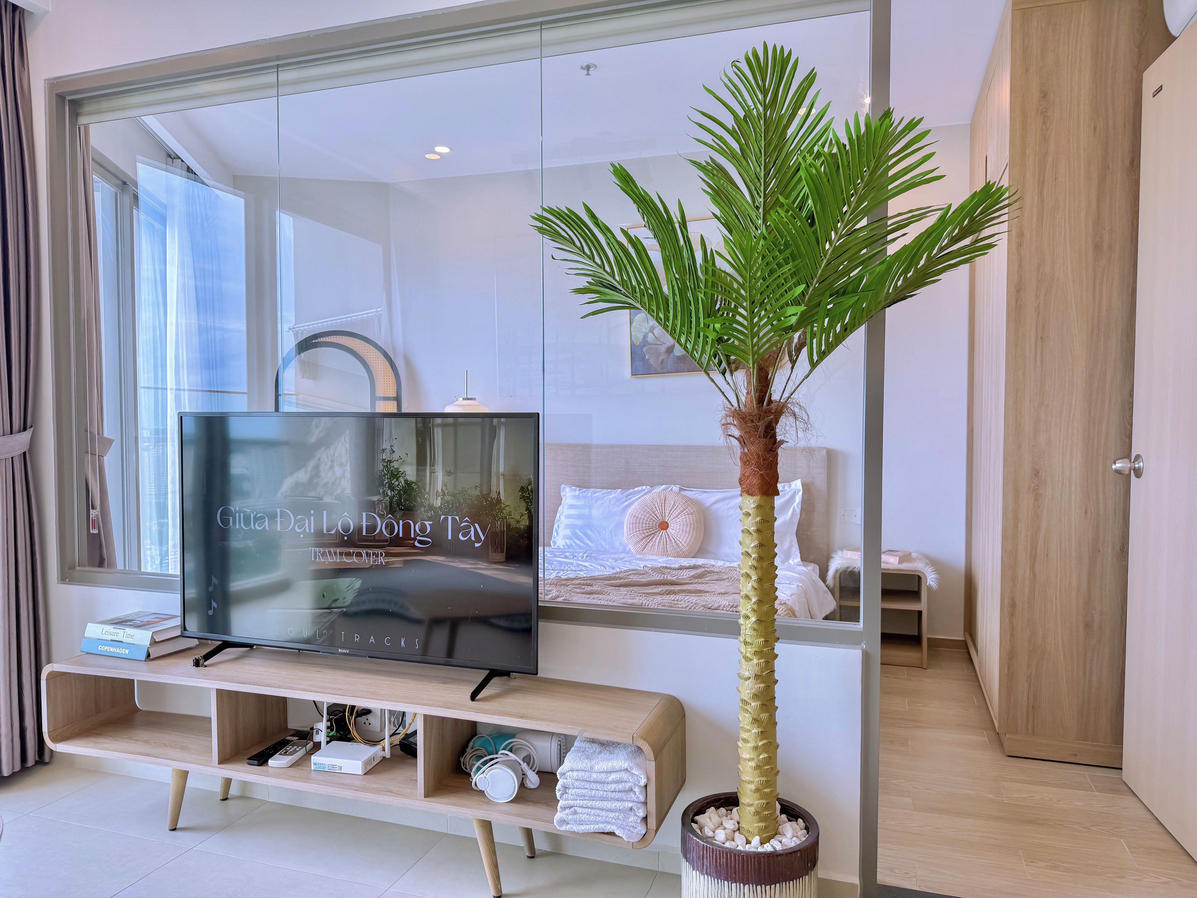 Een moderne hotelkamer met een grote flatscreen televisie op een houten tv-meubel, een bed met wit beddengoed, een kamerplant in de vorm van een palmboom en een houten deur.