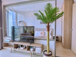 A modern hotel room with a large flat-screen TV on a wooden TV stand, a bed with white bedding, a palm tree plant, and a wooden door. - Lisa House - The Song Condotel Vung Tau  (Ho Chi Minh City)