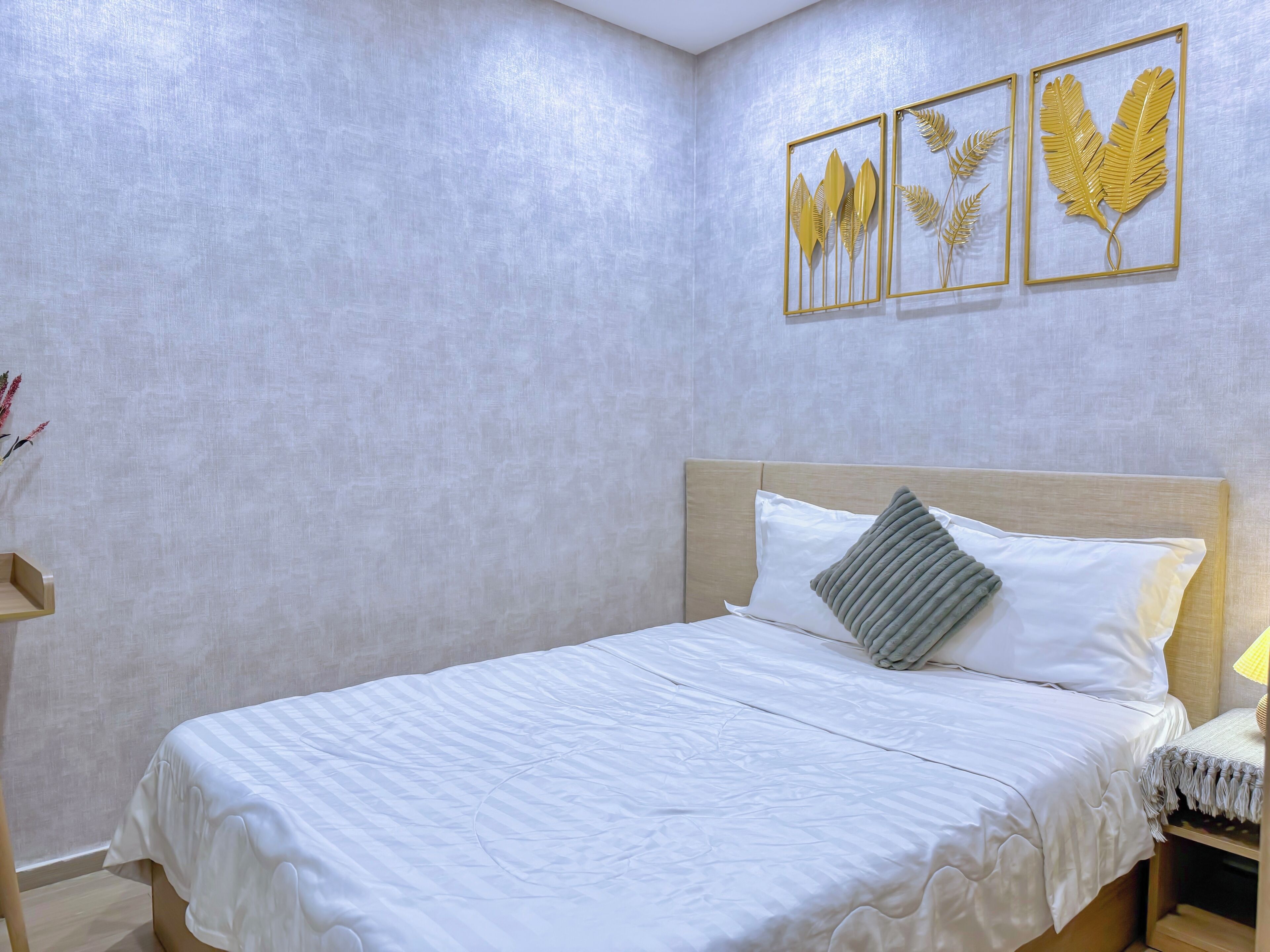 Un letto rifatto con lenzuola bianche e un cuscino, un comodino con una lampada, e una parete con tre decorazioni a foglia dorata incorniciate.