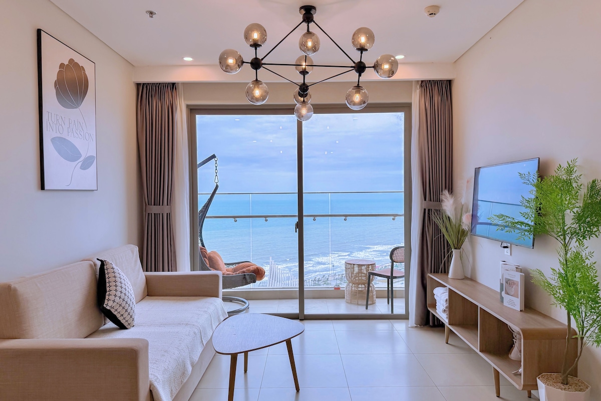 A modern living room with a sofa, a coffee table, and a TV. A large window offers a view of the ocean.