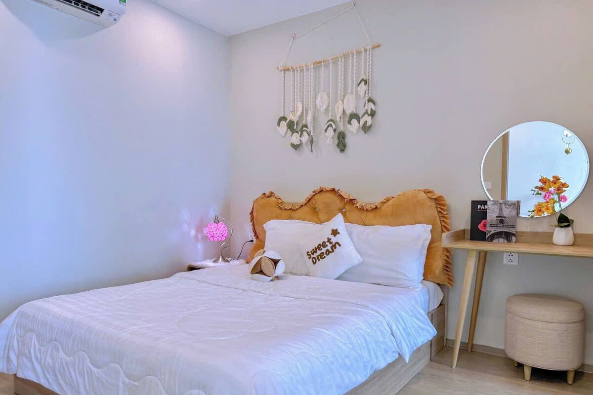A neatly made bed with a white comforter and pillows, a bedside table with a mirror, a decorative wall hanging, and a small stool.