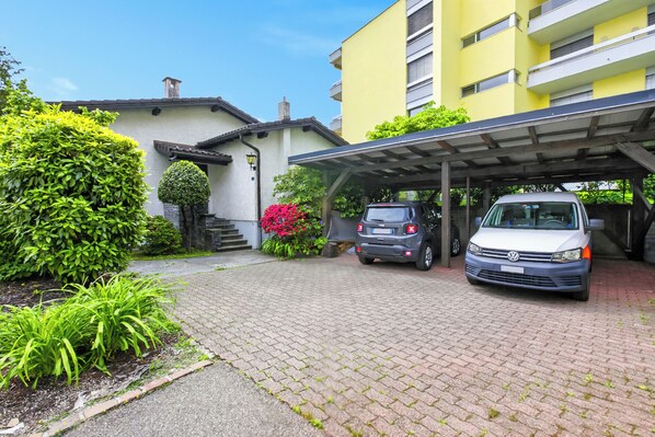 A building with a covered parking area, a white car, and a grey car parked in front. - Panoramic Garden Lake View in Locarno Monti, Locarno, Switzerland (Locarno)