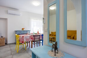 Un comedor con una mesa dispuesta para dos personas, un banco azul con una figurita de caballo y un espejo grande que refleja un sofá amarillo.