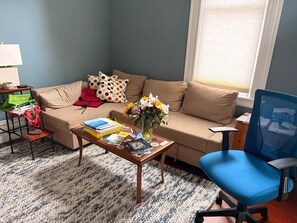 Ein Wohnzimmer mit einer Couch, einem Couchtisch mit Büchern, einer Vase mit Blumen und einem blauen Bürostuhl.