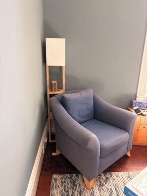 Ein Eckzimmer mit grauem Sessel, einem Holzbodenregal mit Lampe und einem kleinen Tisch mit Gegenständen.