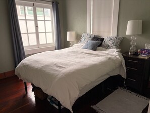 Ein Schlafzimmer mit einem Bett, zwei Nachttischlampen, einem Nachttisch, einem Fenster mit Vorhängen und einem Holzboden.