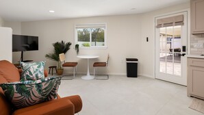 A modern living room with a TV, orange sofa, and tropical pillows. There's a dining area with a table and chairs, a window, and a view of the outdoors. - Pool and Pool House in McAllen (McAllen)